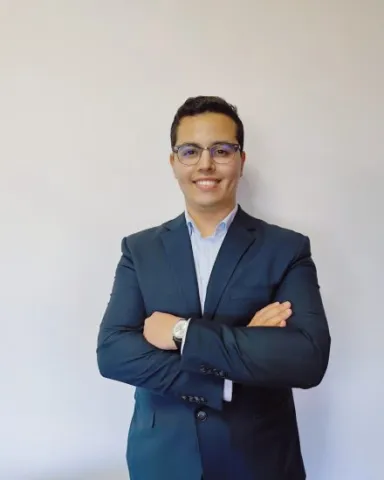 A man in a suit and glasses stands before a white wall, displaying a confident, professional posture.